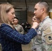 A woman in civilian attire is pinning a medal onto a soldier in U.S. Army OCP (Operational Camouflage Pattern) uniform during what appears to be a formal military awards ceremony. The event is taking place inside a military facility or hangar, with other uniformed personnel and civilians visible in the background. The ceremony appears to be an official recognition or decoration event.