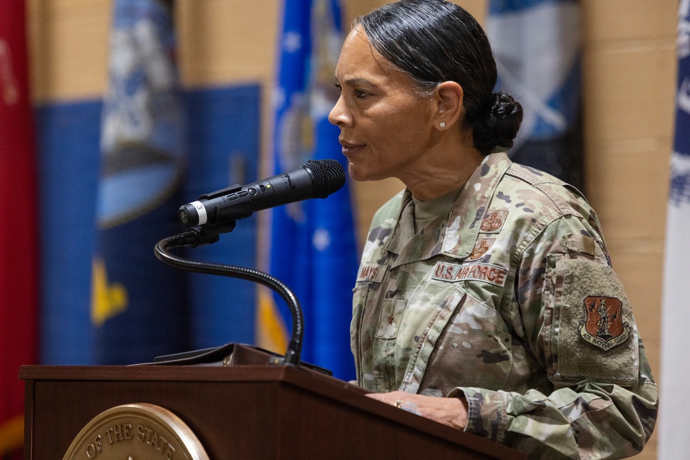 A U.S. Air Force officer, identifiable by the 'U.S. AIR FORCE' tape and 'Air National Guard' shoulder patch, is speaking at a wooden podium equipped with a gooseneck microphone. The officer's name tape reads 'MAYS' and a single star insignia is visible on the uniform, indicating the rank of Brigadier General. The setting appears to be an official military ceremony with flags visible in the background.