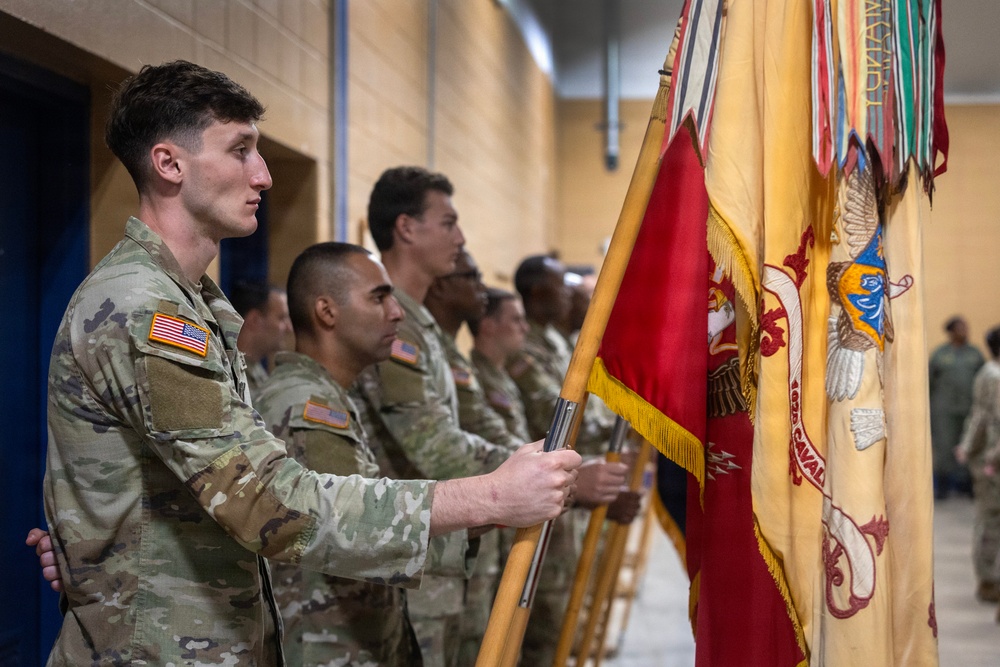 A group of U.S. Army soldiers in OCP (Operational Camouflage Pattern) uniforms stand in a formal line during a military ceremony. The foreground soldier holds multiple unit guidons and flags, including what appears to be a Cavalry unit standard with gold fringe. The setting appears to be an indoor gymnasium or assembly hall.