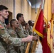 A group of U.S. Army soldiers in OCP (Operational Camouflage Pattern) uniforms stand in a formal line during a military ceremony. The foreground soldier holds multiple unit guidons and flags, including what appears to be a Cavalry unit standard with gold fringe. The setting appears to be an indoor gymnasium or assembly hall.