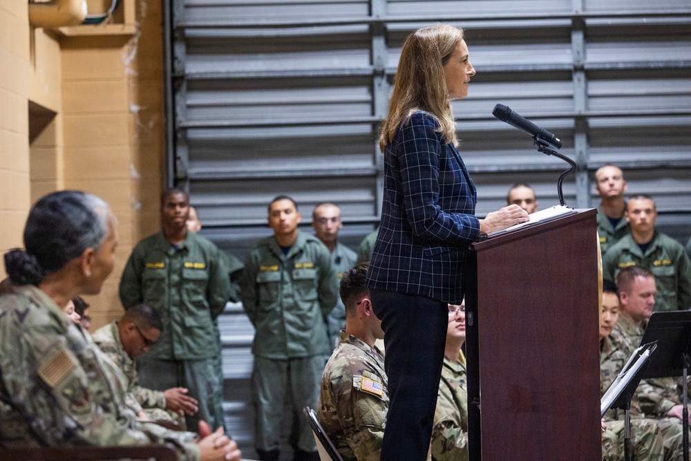 A civilian woman in a navy blue plaid blazer is speaking at a wooden podium equipped with a microphone, addressing a military audience. The audience includes personnel in U.S. Army combat uniforms (ACU/OCP pattern) seated in the foreground, while a row of personnel in olive drab flight suits stand at attention in the background. The setting appears to be a military hangar or warehouse facility.
