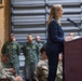 A civilian woman in a navy blue plaid blazer is speaking at a wooden podium equipped with a microphone, addressing a military audience. The audience includes personnel in U.S. Army combat uniforms (ACU/OCP pattern) seated in the foreground, while a row of personnel in olive drab flight suits stand at attention in the background. The setting appears to be a military hangar or warehouse facility.