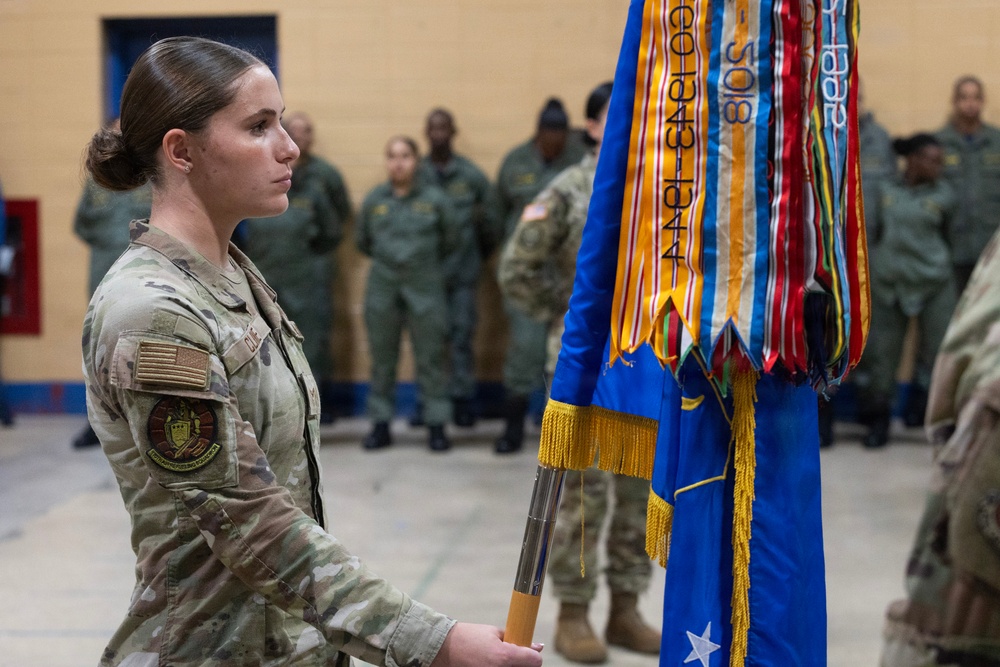 A female military service member in OCP (Operational Camouflage Pattern) uniform holds a guidon or unit colors staff during what appears to be a formal military ceremony held indoors in a gymnasium or drill hall. The flag displayed is blue with gold fringe, consistent with a U.S. Air Force unit colors, adorned with multiple campaign and award streamers. Additional uniformed personnel stand in formation in the background.