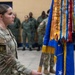 A female military service member in OCP (Operational Camouflage Pattern) uniform holds a guidon or unit colors staff during what appears to be a formal military ceremony held indoors in a gymnasium or drill hall. The flag displayed is blue with gold fringe, consistent with a U.S. Air Force unit colors, adorned with multiple campaign and award streamers. Additional uniformed personnel stand in formation in the background.