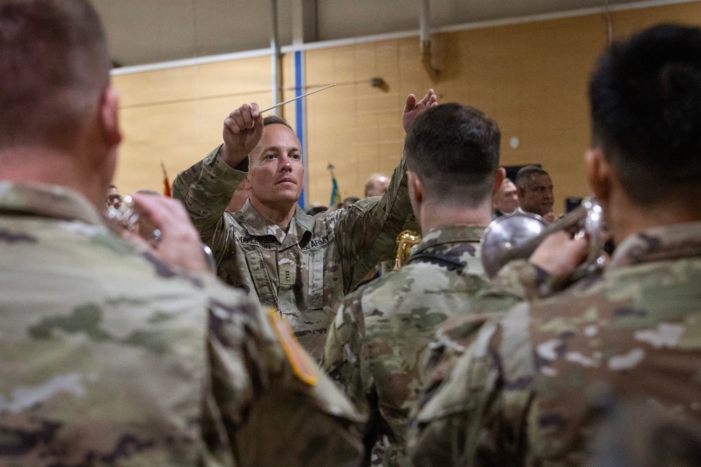 A U.S. Army soldier in OCP (Operational Camouflage Pattern) uniform is conducting a military band, holding a conductor's baton with both hands raised. The scene takes place indoors, likely in a gymnasium or assembly hall, with other uniformed soldiers visible as band members in the foreground and background. Musical instruments including what appears to be a tuba or sousaphone are visible among the performers.