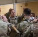 A U.S. Army soldier in OCP (Operational Camouflage Pattern) uniform is conducting a military band, holding a conductor's baton with both hands raised. The scene takes place indoors, likely in a gymnasium or assembly hall, with other uniformed soldiers visible as band members in the foreground and background. Musical instruments including what appears to be a tuba or sousaphone are visible among the performers.