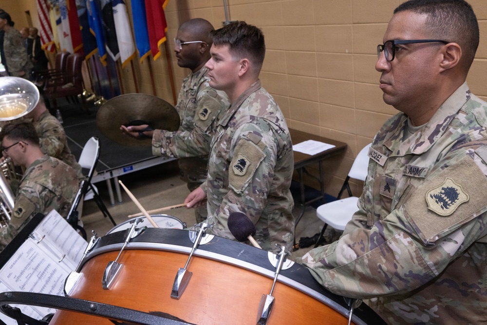 U.S. Army soldiers in OCP (Operational Camouflage Pattern) uniforms perform as part of a military band during what appears to be an official ceremony or event. The percussion section is prominently featured, with soldiers playing snare drum, bass drum, and cymbals. Military flags and other band members with brass instruments are visible in the background.