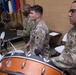 U.S. Army soldiers in OCP (Operational Camouflage Pattern) uniforms perform as part of a military band during what appears to be an official ceremony or event. The percussion section is prominently featured, with soldiers playing snare drum, bass drum, and cymbals. Military flags and other band members with brass instruments are visible in the background.