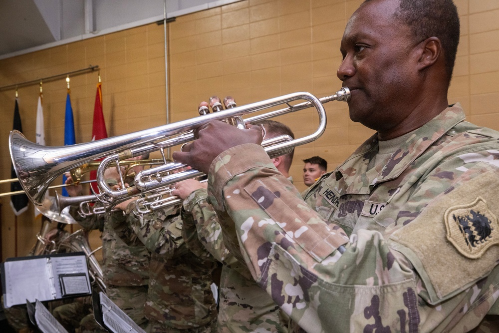 U.S. Army soldiers in MultiCam/OCP camouflage uniforms are performing as a military band, playing silver trumpets during what appears to be an official ceremony or event. The foreground soldier, whose name tape reads 'HEND' (partially visible), is prominently playing a silver trumpet. The setting appears to be an indoor gymnasium or assembly hall with military flags visible in the background.