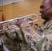 U.S. Army soldiers in MultiCam/OCP camouflage uniforms are performing as a military band, playing silver trumpets during what appears to be an official ceremony or event. The foreground soldier, whose name tape reads 'HEND' (partially visible), is prominently playing a silver trumpet. The setting appears to be an indoor gymnasium or assembly hall with military flags visible in the background.