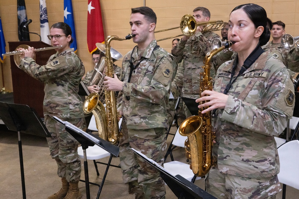 A U.S. Army military band is performing at an indoor ceremony or official event. Soldiers in OCP (Operational Camouflage Pattern) uniforms are playing various instruments including flute, baritone saxophone, alto saxophone, trombone, and other brass instruments. Multiple flags and a podium with a microphone are visible in the background, indicating a formal military function.