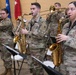 A U.S. Army military band is performing at an indoor ceremony or official event. Soldiers in OCP (Operational Camouflage Pattern) uniforms are playing various instruments including flute, baritone saxophone, alto saxophone, trombone, and other brass instruments. Multiple flags and a podium with a microphone are visible in the background, indicating a formal military function.