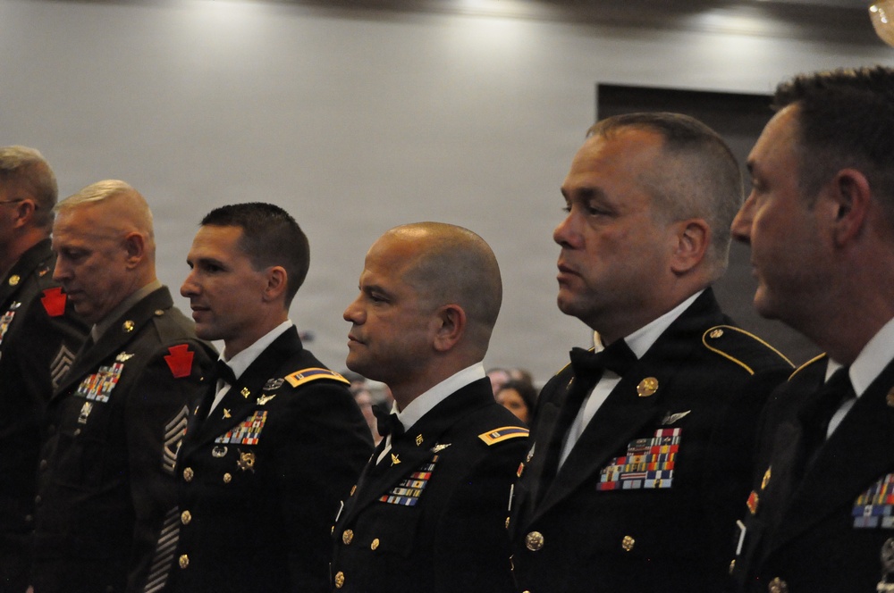 A group of U.S. Army personnel in dress uniforms (Army Service Uniform/Mess Dress) are standing in a formal indoor setting, likely at a military ball or formal ceremony. The soldiers display numerous medals, ribbons, and insignia on their uniforms. They appear to be standing at attention or in a formal posture facing a common direction.