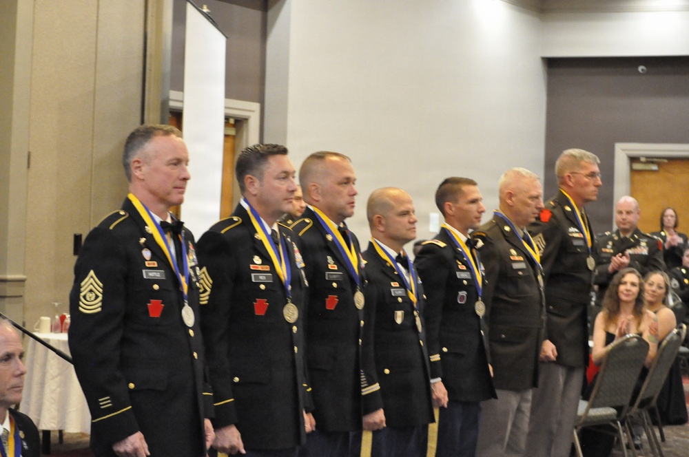 A group of military personnel in dress uniforms are standing in a formal line at what appears to be an awards ceremony or military banquet. Each individual is wearing a medal on a blue and yellow ribbon around their neck, along with various rank insignia and unit patches. An audience is visible in the background, seated at tables in what appears to be a hotel ballroom or conference facility.