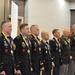 A group of military personnel in dress uniforms are standing in a formal line at what appears to be an awards ceremony or military banquet. Each individual is wearing a medal on a blue and yellow ribbon around their neck, along with various rank insignia and unit patches. An audience is visible in the background, seated at tables in what appears to be a hotel ballroom or conference facility.