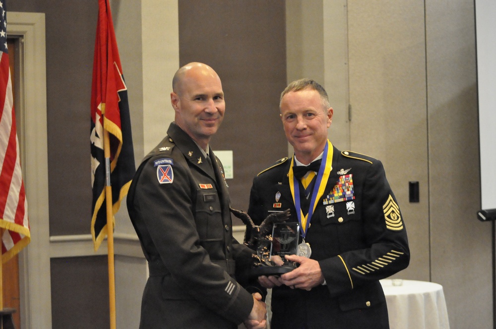 Two U.S. Army soldiers pose together at a formal military ceremony, jointly holding a trophy or award. The soldier on the right is wearing dress blues with an extensive rack of ribbons and medals, including a medallion on a blue and yellow ribbon around his neck. The soldier on the left is in Army Service Uniform (ASU greens) with a 10th Mountain Division patch visible.