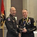 Two U.S. Army soldiers pose together at a formal military ceremony, jointly holding a trophy or award. The soldier on the right is wearing dress blues with an extensive rack of ribbons and medals, including a medallion on a blue and yellow ribbon around his neck. The soldier on the left is in Army Service Uniform (ASU greens) with a 10th Mountain Division patch visible.