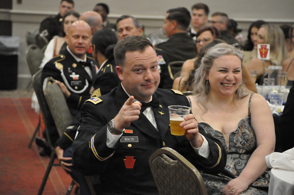 A formal military ball or gala event featuring uniformed U.S. Army personnel and civilian guests seated at banquet tables. The foreground shows a soldier identified by nameplate as 'BOLTZ' in dress uniform pointing at the camera while holding a beer, seated next to a laughing woman in a formal gown. The background shows a crowded banquet hall with numerous attendees in military dress uniforms and formal civilian attire.