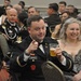 A formal military ball or gala event featuring uniformed U.S. Army personnel and civilian guests seated at banquet tables. The foreground shows a soldier identified by nameplate as 'BOLTZ' in dress uniform pointing at the camera while holding a beer, seated next to a laughing woman in a formal gown. The background shows a crowded banquet hall with numerous attendees in military dress uniforms and formal civilian attire.