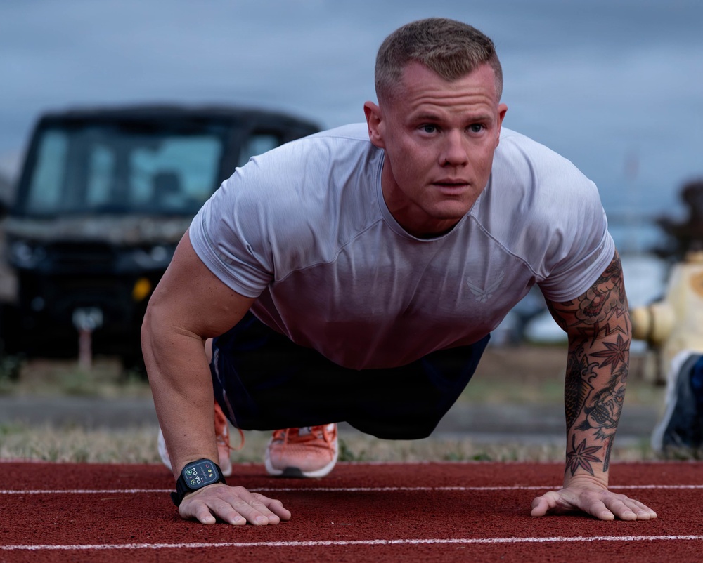 A muscular male athlete performs push-ups on a red outdoor running track. He is wearing a white athletic t-shirt, dark shorts, orange sneakers, and a smartwatch on his left wrist. His right forearm and arm are covered in visible tattoos.