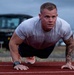 A muscular male athlete performs push-ups on a red outdoor running track. He is wearing a white athletic t-shirt, dark shorts, orange sneakers, and a smartwatch on his left wrist. His right forearm and arm are covered in visible tattoos.