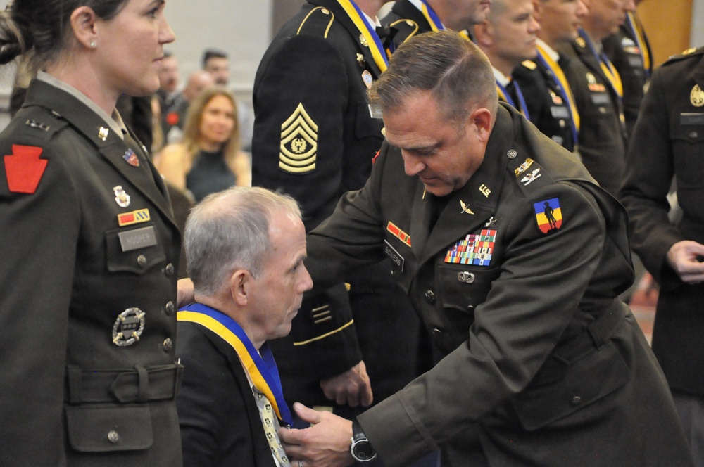 A U.S. Army military ceremony is taking place in which a senior officer is placing or adjusting a medal/medallion with a blue and gold ribbon around the neck of a seated civilian or recipient. Multiple uniformed Army personnel are present in the background, many already wearing similar blue and gold medallions. The setting appears to be an indoor venue consistent with a formal military awards or induction ceremony.