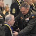 A U.S. Army military ceremony is taking place in which a senior officer is placing or adjusting a medal/medallion with a blue and gold ribbon around the neck of a seated civilian or recipient. Multiple uniformed Army personnel are present in the background, many already wearing similar blue and gold medallions. The setting appears to be an indoor venue consistent with a formal military awards or induction ceremony.