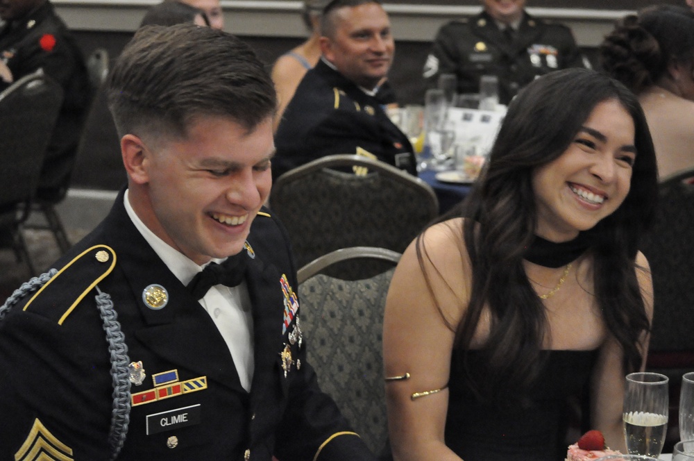A U.S. Army soldier in dress uniform (Army Service Uniform with bow tie) and a woman in a black dress are seated at a table, both laughing and smiling. The setting appears to be a formal military ball or banquet event. Additional soldiers in dress uniforms are visible at tables in the background.
