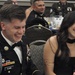 A U.S. Army soldier in dress uniform (Army Service Uniform with bow tie) and a woman in a black dress are seated at a table, both laughing and smiling. The setting appears to be a formal military ball or banquet event. Additional soldiers in dress uniforms are visible at tables in the background.