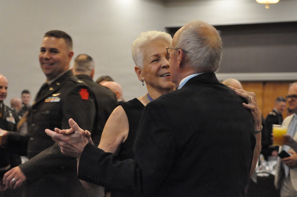 A formal military event or banquet is taking place in a large indoor venue. Two older civilians, a woman with short white hair and a man in a dark suit, are embracing or dancing in the foreground. Military personnel in Army dress uniforms are visible in the background, along with other attendees.