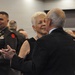 A formal military event or banquet is taking place in a large indoor venue. Two older civilians, a woman with short white hair and a man in a dark suit, are embracing or dancing in the foreground. Military personnel in Army dress uniforms are visible in the background, along with other attendees.