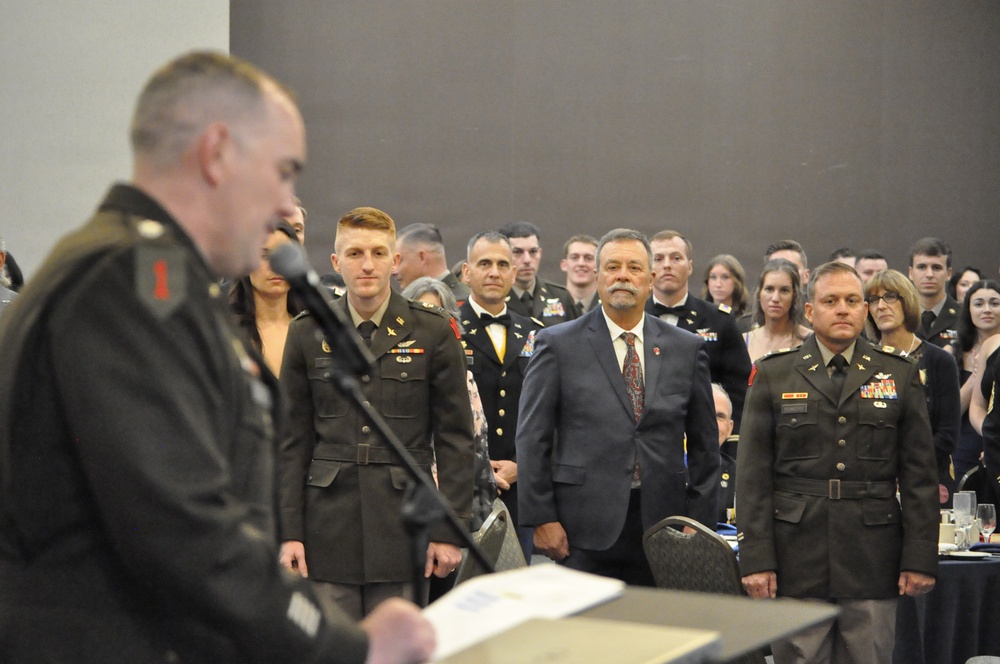 A formal military ceremony or banquet is taking place in a large indoor venue. A uniformed officer stands at a podium with a microphone, addressing a crowd of military personnel in dress uniforms and civilians standing at attention. The event appears to be an awards ceremony, formal dinner, or recognition event based on the attire and setting.