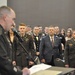 A formal military ceremony or banquet is taking place in a large indoor venue. A uniformed officer stands at a podium with a microphone, addressing a crowd of military personnel in dress uniforms and civilians standing at attention. The event appears to be an awards ceremony, formal dinner, or recognition event based on the attire and setting.