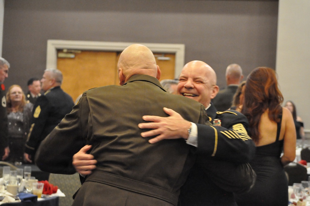 Two bald men in military dress uniforms are embracing warmly at what appears to be a formal military banquet or ceremony. The man facing the camera is smiling broadly, suggesting a joyful reunion or celebratory moment. The setting is a banquet hall with other uniformed personnel and civilians visible in the background.