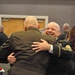Two bald men in military dress uniforms are embracing warmly at what appears to be a formal military banquet or ceremony. The man facing the camera is smiling broadly, suggesting a joyful reunion or celebratory moment. The setting is a banquet hall with other uniformed personnel and civilians visible in the background.