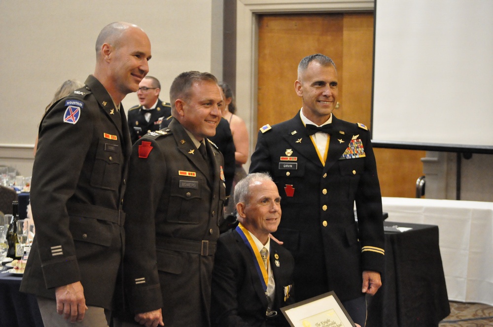 A formal military ceremony or banquet featuring three U.S. Army officers in uniform posing with a seated civilian who is wearing a medallion/award around his neck and holding a framed certificate. The setting appears to be a banquet hall with tables and other attendees visible in the background. This appears to be an awards or recognition ceremony.