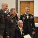 A formal military ceremony or banquet featuring three U.S. Army officers in uniform posing with a seated civilian who is wearing a medallion/award around his neck and holding a framed certificate. The setting appears to be a banquet hall with tables and other attendees visible in the background. This appears to be an awards or recognition ceremony.