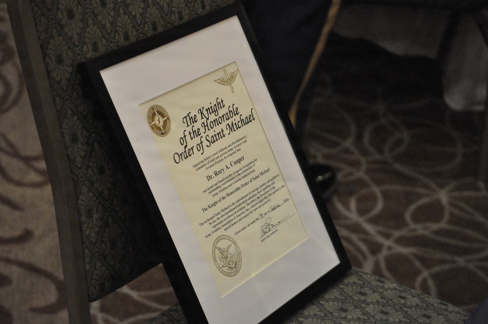 A framed certificate titled 'The Knight of the Honorable Order of Saint Michael' is displayed on an easel or stand. The certificate is awarded to Dr. Rory A. Cooper and is associated with Army Aviation. It features official seals, decorative elements, and a signature at the bottom.