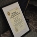 A framed certificate titled 'The Knight of the Honorable Order of Saint Michael' is displayed on an easel or stand. The certificate is awarded to Dr. Rory A. Cooper and is associated with Army Aviation. It features official seals, decorative elements, and a signature at the bottom.