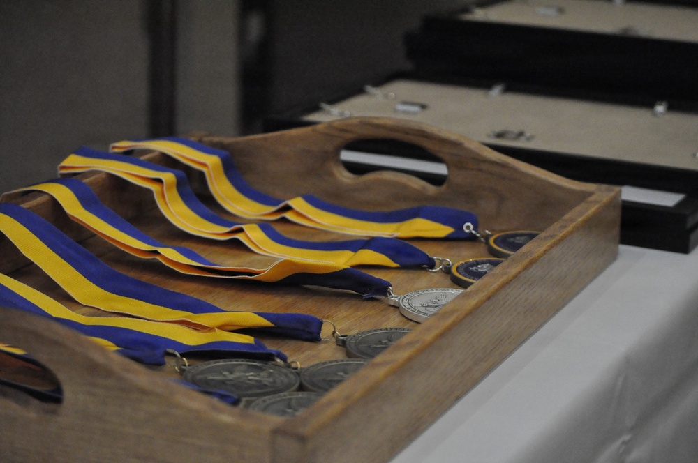 A wooden carrying tray holds multiple medals with blue and yellow striped ribbons, arranged on a white-cloth-covered table. The medals appear to be silver-toned and are laid out in preparation for an awards ceremony or presentation. Additional display cases or trays are visible in the blurred background.