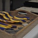 A wooden carrying tray holds multiple medals with blue and yellow striped ribbons, arranged on a white-cloth-covered table. The medals appear to be silver-toned and are laid out in preparation for an awards ceremony or presentation. Additional display cases or trays are visible in the blurred background.