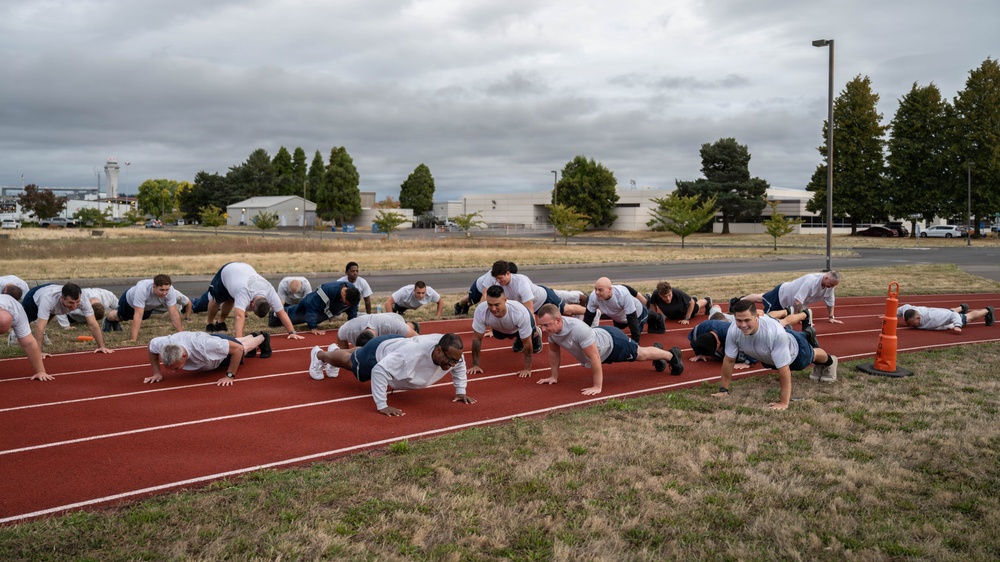 A large group of individuals in matching white t-shirts and dark shorts are performing push-ups on a red outdoor running track. The setting appears to be a military or institutional fitness facility, with buildings, trees, and an overcast sky visible in the background. The group is organized in multiple rows, suggesting a structured physical training session.