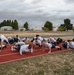 A large group of individuals in matching white t-shirts and dark shorts are performing push-ups on a red outdoor running track. The setting appears to be a military or institutional fitness facility, with buildings, trees, and an overcast sky visible in the background. The group is organized in multiple rows, suggesting a structured physical training session.