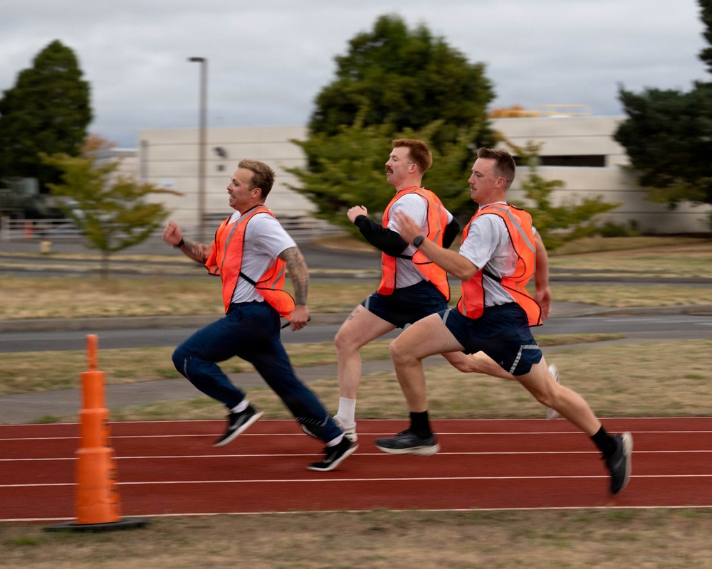 Three men in matching uniforms consisting of white t-shirts, orange reflective safety vests, and dark shorts or pants are running on a red athletic track. A fourth runner is partially visible in the background. The setting appears to be a military or institutional facility based on the buildings and infrastructure visible in the background.