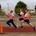 Three men in matching uniforms consisting of white t-shirts, orange reflective safety vests, and dark shorts or pants are running on a red athletic track. A fourth runner is partially visible in the background. The setting appears to be a military or institutional facility based on the buildings and infrastructure visible in the background.