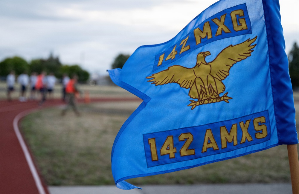 A blue military unit guidon flag for the 142nd Aircraft Maintenance Squadron (142 AMXS) under the 14Z Maintenance Group (14Z MXG) is prominently displayed in the foreground. The flag features a gold eagle emblem and gold lettering on a blue background. In the blurred background, personnel in athletic attire are gathered near a running track, suggesting a physical fitness event.