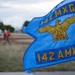 A blue military unit guidon flag for the 142nd Aircraft Maintenance Squadron (142 AMXS) under the 14Z Maintenance Group (14Z MXG) is prominently displayed in the foreground. The flag features a gold eagle emblem and gold lettering on a blue background. In the blurred background, personnel in athletic attire are gathered near a running track, suggesting a physical fitness event.