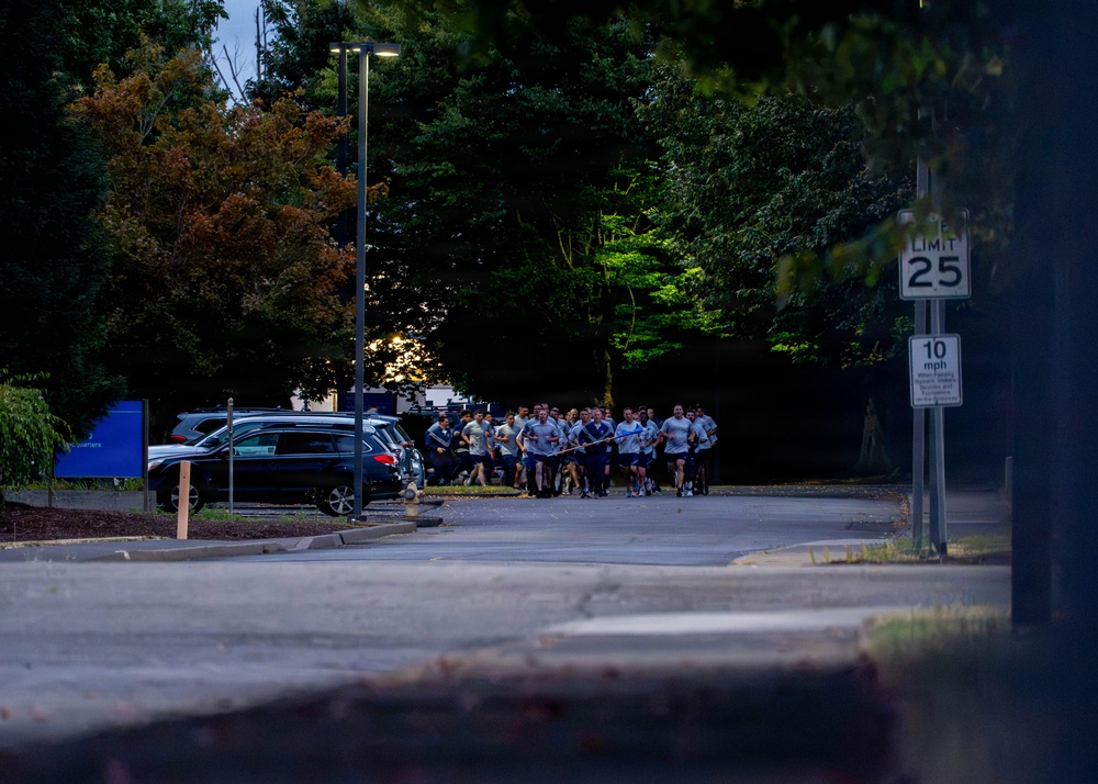 A group of people in matching light-colored athletic shirts are running together in formation along a road or parking area during early morning or dusk conditions. The group appears to be conducting an organized group run, possibly military or institutional physical training. The scene is set in a wooded area with parked vehicles and street signage visible.