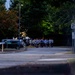 A group of people in matching light-colored athletic shirts are running together in formation along a road or parking area during early morning or dusk conditions. The group appears to be conducting an organized group run, possibly military or institutional physical training. The scene is set in a wooded area with parked vehicles and street signage visible.