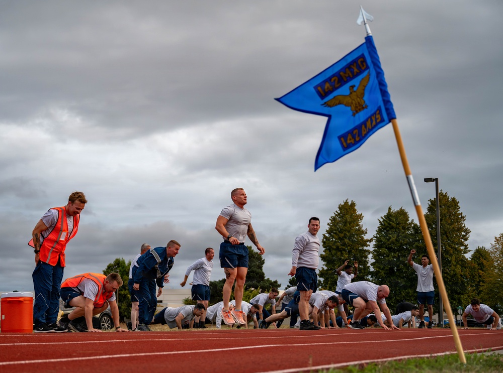A group of military personnel are conducting a physical fitness test or training exercise on an outdoor running track. A blue unit guidon flag marked '142 MXG / 142 AMXS' is prominently displayed in the foreground, indicating this is a U.S. Air National Guard unit event. Participants are performing various exercises including push-ups and running, while two individuals in orange safety vests appear to be officiating or monitoring the event.